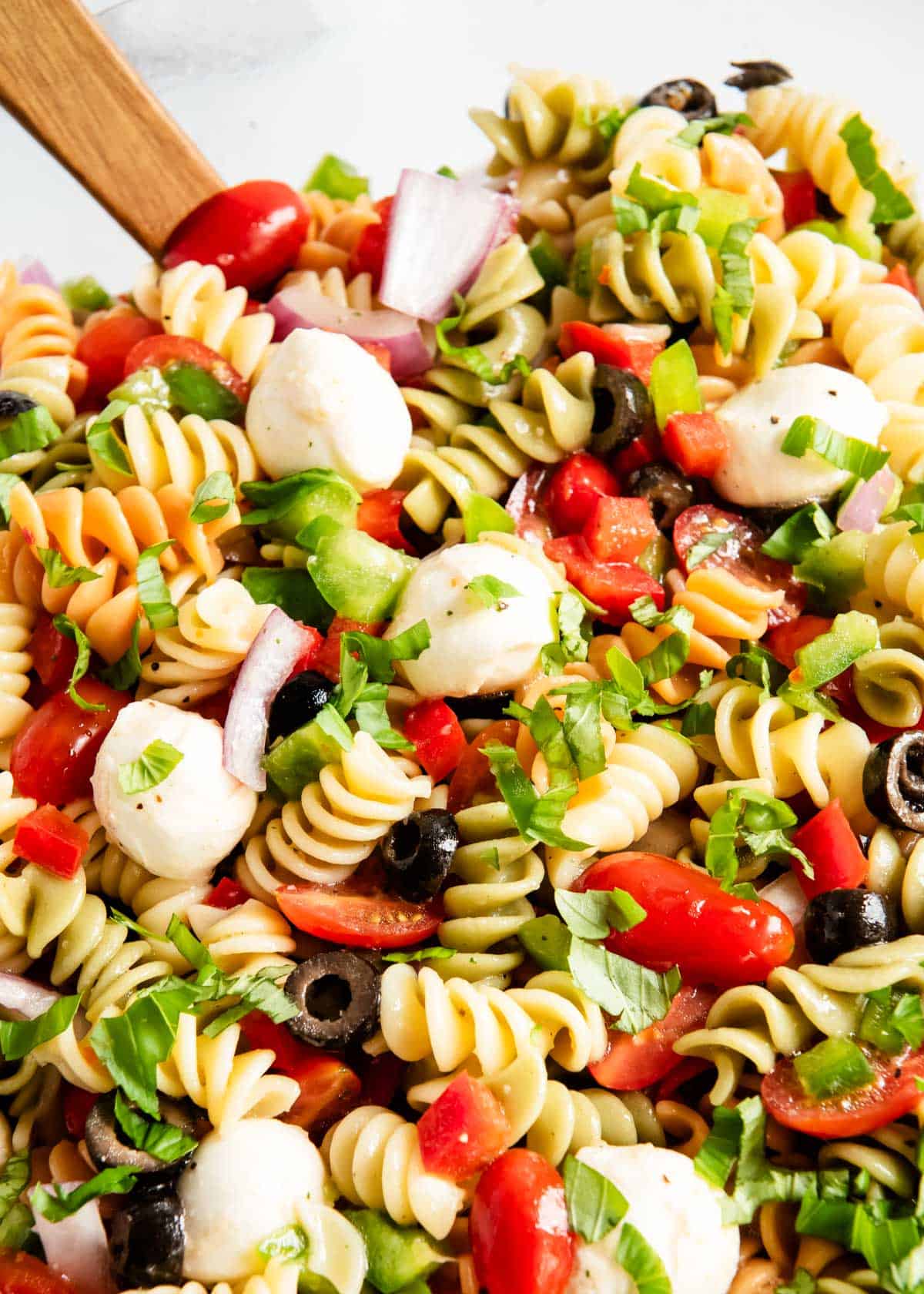 Tossing pasta salad ingredients with Italian dressing in a large glass bowl using wooden salad servers.